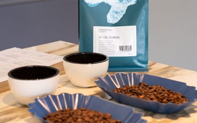 Kaffeebohnen in blauer Schale auf einem Tisch, dahinter zwei weiße Tassen und Kaffeeverpackung