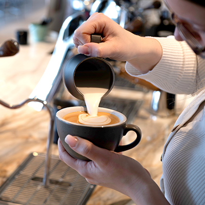 Aufgeschäumte Milch wird mit einem Milchkännchen auf den Espresso in der Cappuccinotasse gegossen.